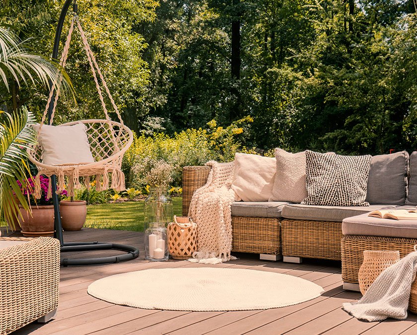 Backyard hardwood deck with a beige carpet, rattan couch, mesh swing, pillows and blanket, and trees in the background.