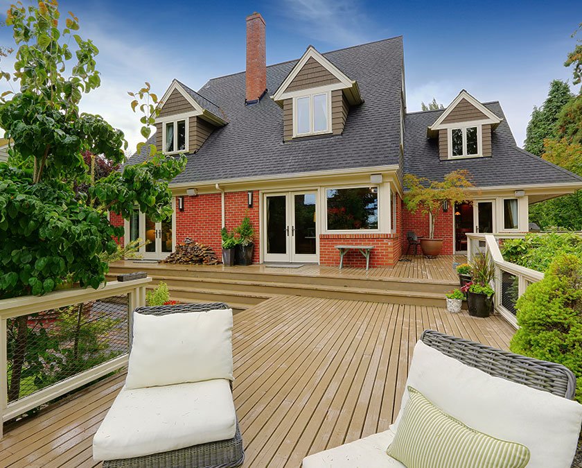Large deck in the backyard of a house on a rainy day, with rattan chairs, pillows, and a tree nearby.