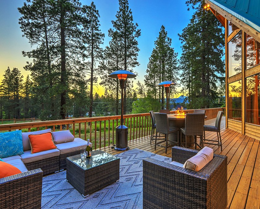 Sunset view of stylish hardwood deck, with rattan couch and chairs, blue and orange pillows, and tall trees in the background.