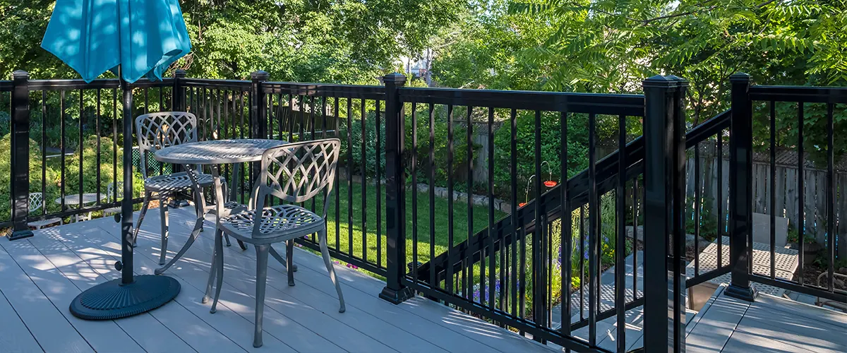 A wrought iron railing on a deck with a plastic table, two chairs, and a blue umbrella