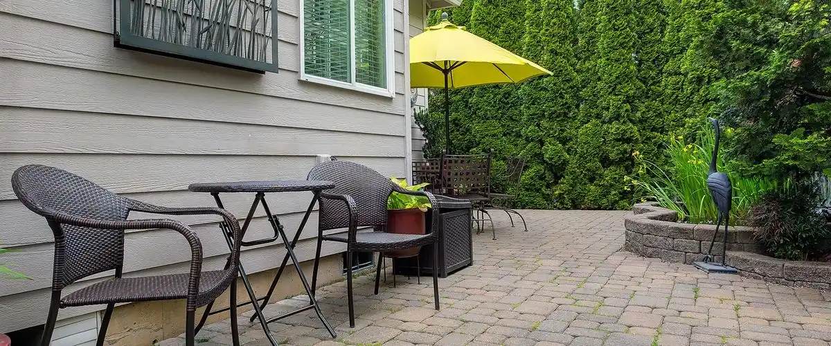 How to Build a Paver Patio DIY: 5 Simple Steps A completed cobblestone paver patio next to a light gray house, featuring brown woven outdoor furniture, a small folding table, a bright yellow umbrella, and a lush green hedge backdrop used for privacy