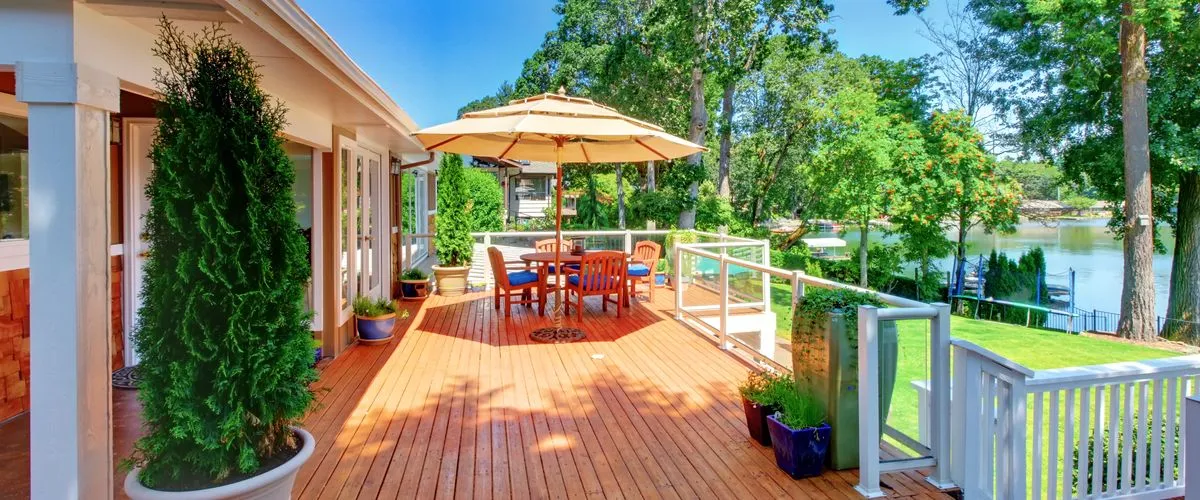 The Top Deck Trends for 2026 Sunny wooden deck with patio furniture, a large umbrella, and a serene lake view surrounded by green trees.
