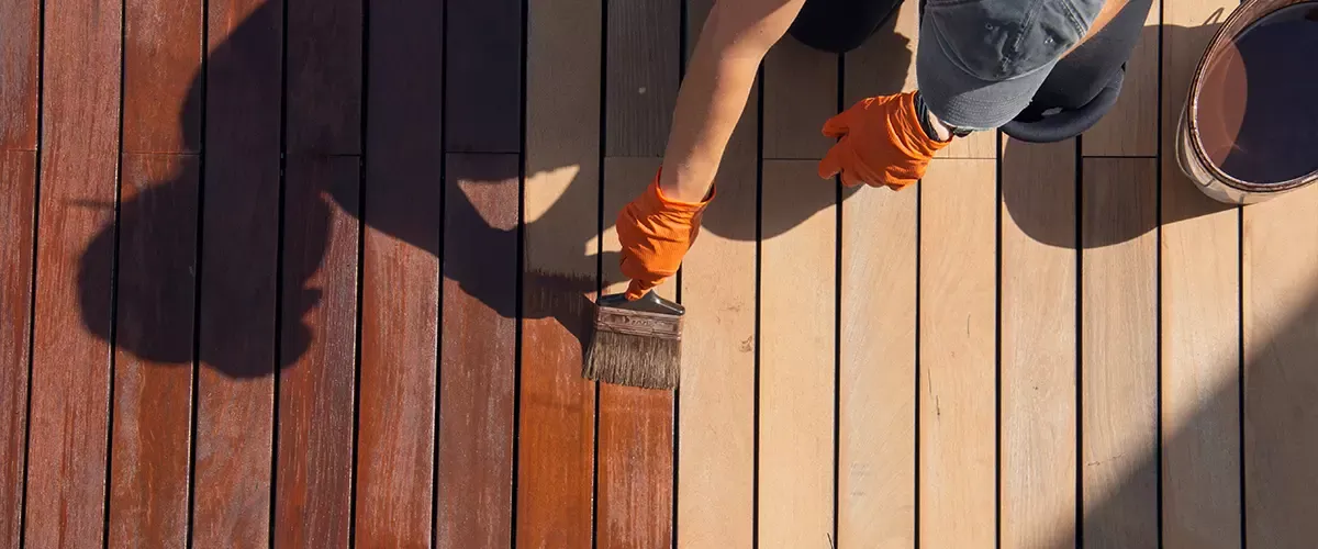 Overhead view of a person wearing orange gloves using a wide brush to apply a dark wood stain to a light-colored timber deck, showing the clear contrast between the treated and untreated wood planks.