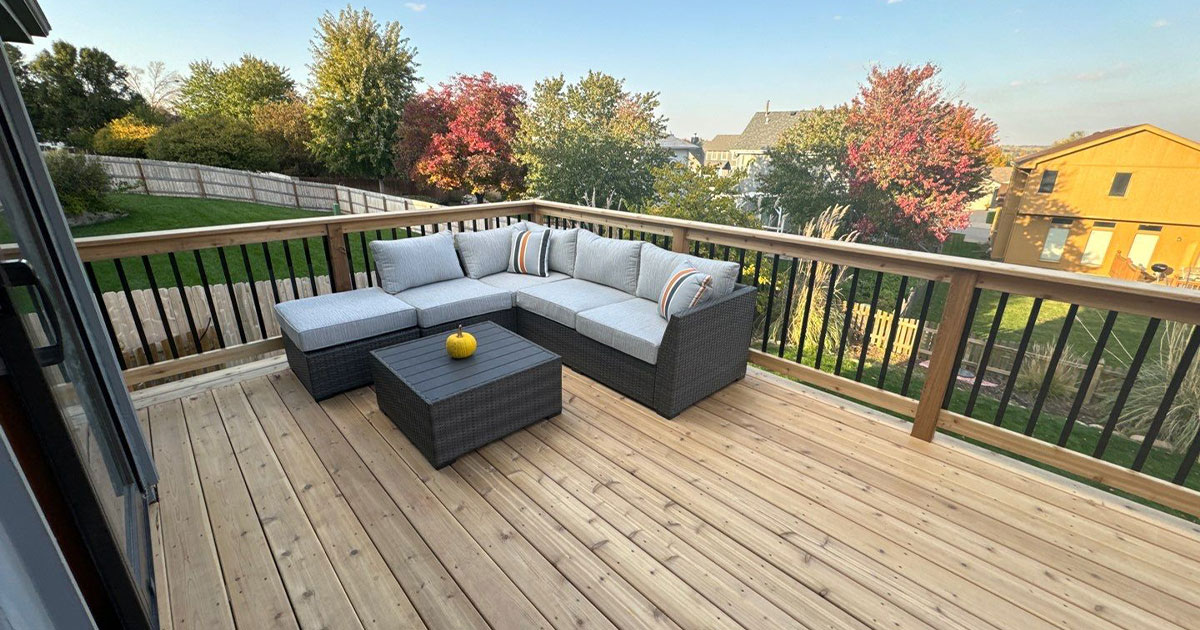 Outdoor patio with sectional sofa and yellow accent pillow on wooden deck, residential backyard view.