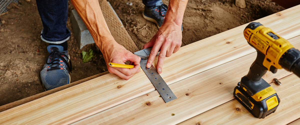 Close-up of a person measuring and marking wooden planks with a ruler and pencil, alongside a yellow power drill, during a deck building project in Bennington.