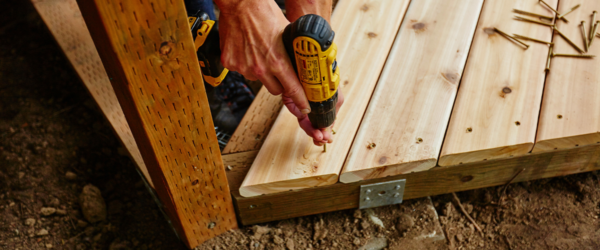 Person using a yellow power drill to screw wooden planks into a deck frame during deck repair, with visible screws and a measuring tool on the side.