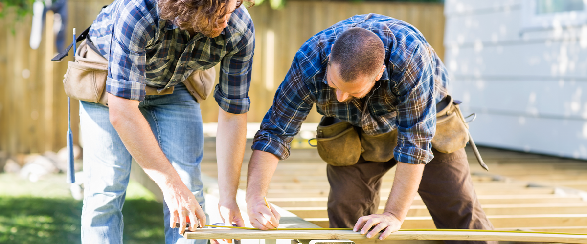 Two men wearing tool belts and plaid shirts working together on a deck installation project, measuring and cutting wood, showcasing teamwork and carpentry skills.