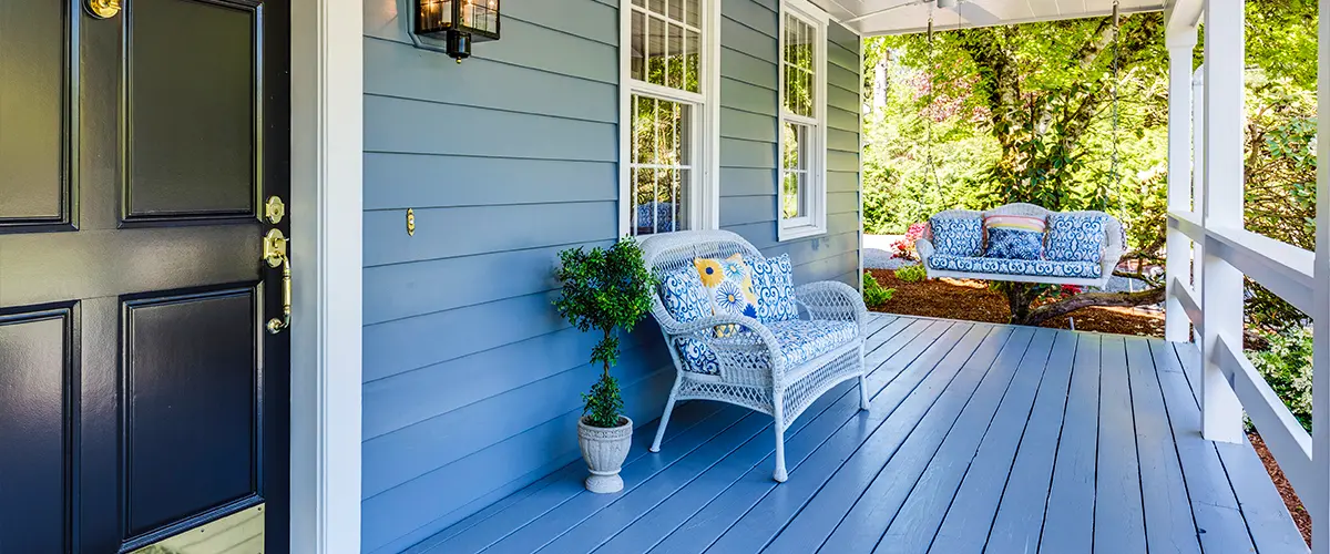 A beautifully designed front porch with blue siding, white trim, and a black front door. Wicker furniture and a swing enhance the cozy ambiance.