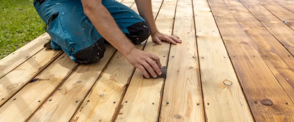 A person sanding a wooden deck with a sanding block, transitioning from old to freshly refinished wood