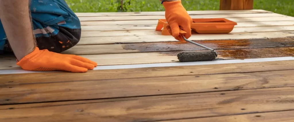 A person wearing orange gloves applying wood stain to a deck with a roller brush, showing a contrast between treated and untreated wood