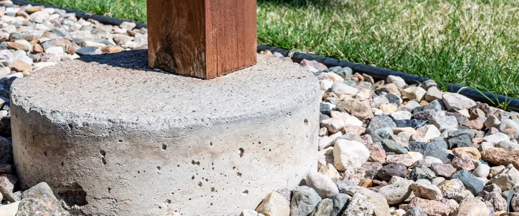 Close-up of a wooden post anchored in a concrete footing with decorative gravel for deck foundation support.