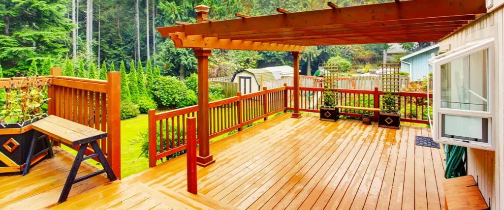 Spacious wooden deck with pergola and built-in bench overlooking a vibrant backyard garden with tall trees.