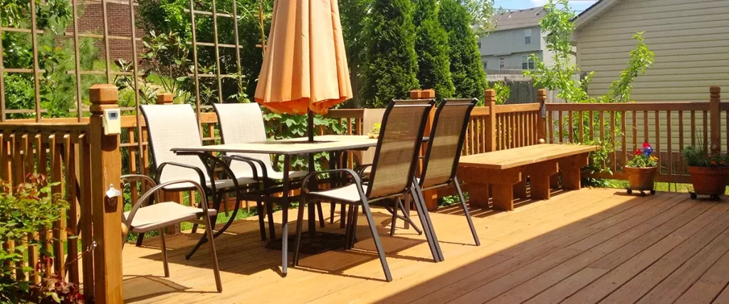 Wooden backyard deck with patio chairs, table, and umbrella surrounded by lush greenery and privacy fence.