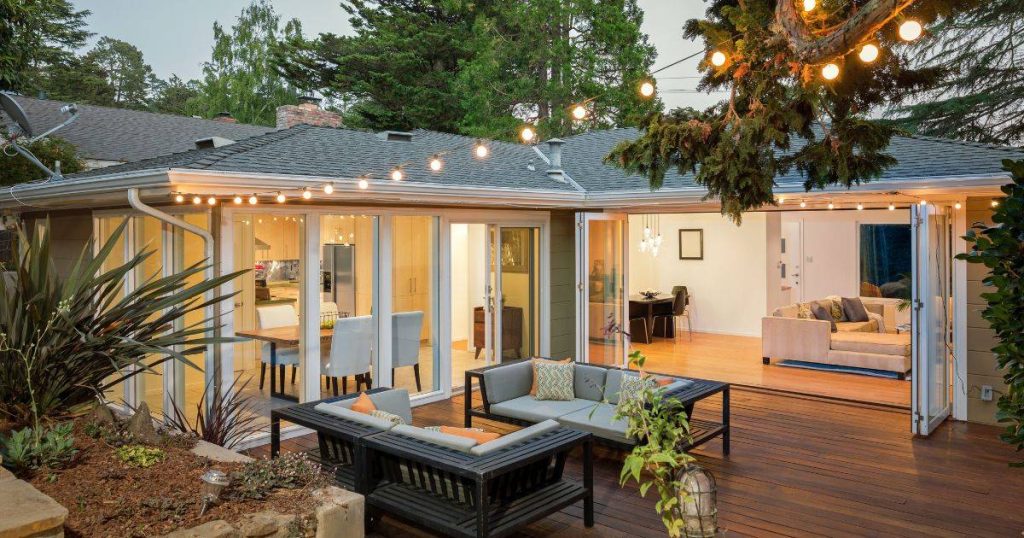 A cozy backyard patio setup with outdoor seating, a fire pit, string lights overhead, and a house with large windows in the background.