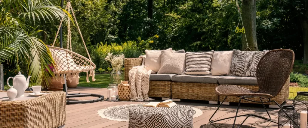 A cozy outdoor patio scene with wicker furniture, layered cushions, blankets, and greenery in the background.