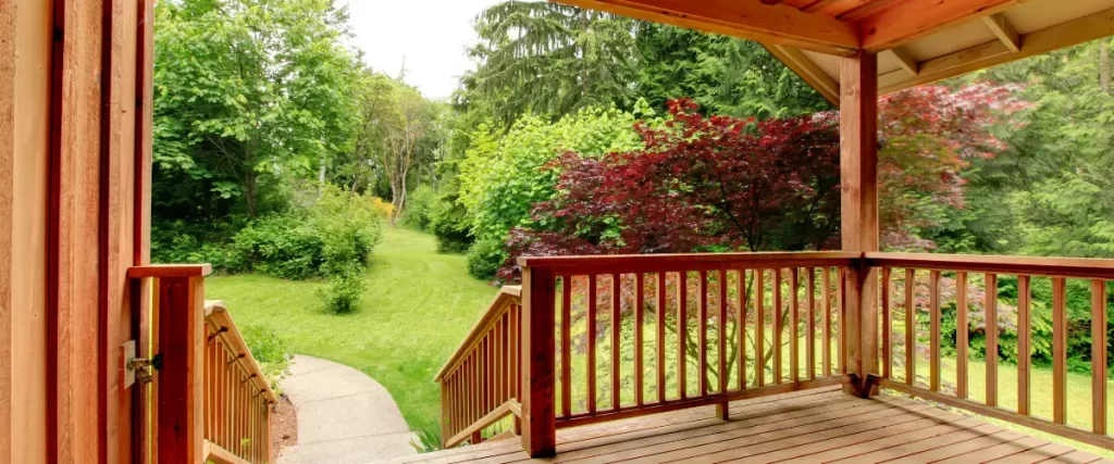 How Omaha’s High Humidity Affects Wooden Decks & the Best Ways to Prevent Rot A small wooden deck with red railings, adjacent to a grassy backyard with trees and shrubs.