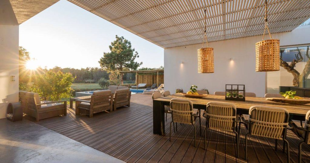 An outdoor dining area on a wooden deck under a slatted pergola roof attached to a modern house.