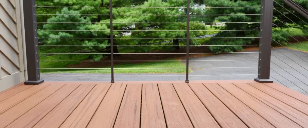 A close-up of a reddish-brown composite deck with modern horizontal cable railing, with trees and a road in the background.