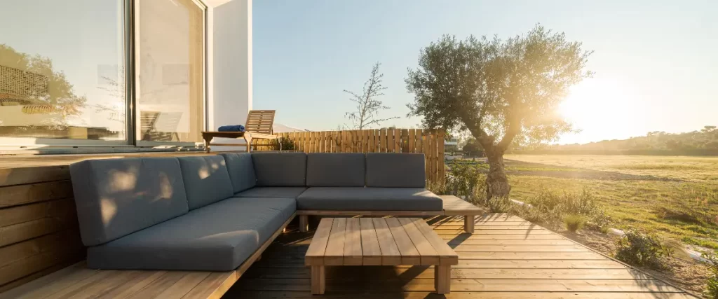 An outdoor sectional sofa on a wooden deck with a low coffee table, set against a sunny, landscaped background.