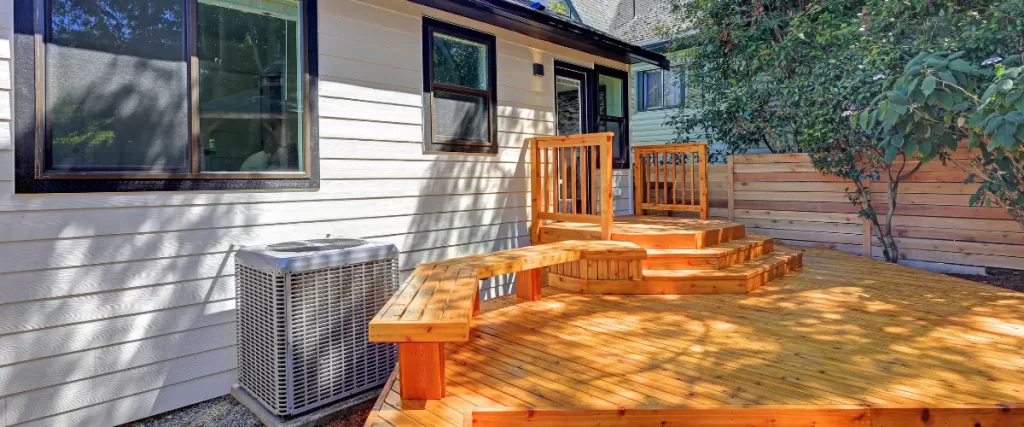 A small wooden deck attached to a house, featuring built-in benches and wooden railings, lit by bright sunlight.