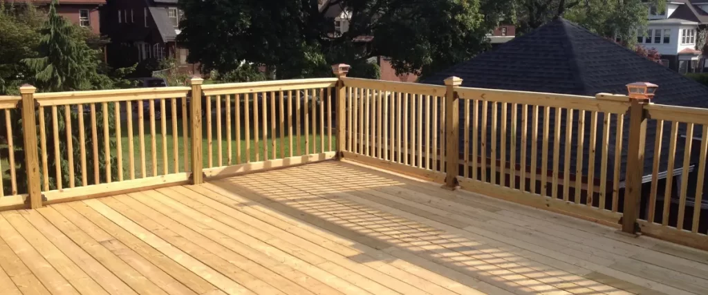 A light-colored wooden deck with vertical wooden railings, casting long shadows in the sunlight.

