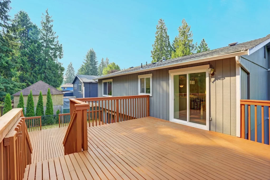 A wooden deck attached to a single-story house, with a railing and surrounded by trees.


