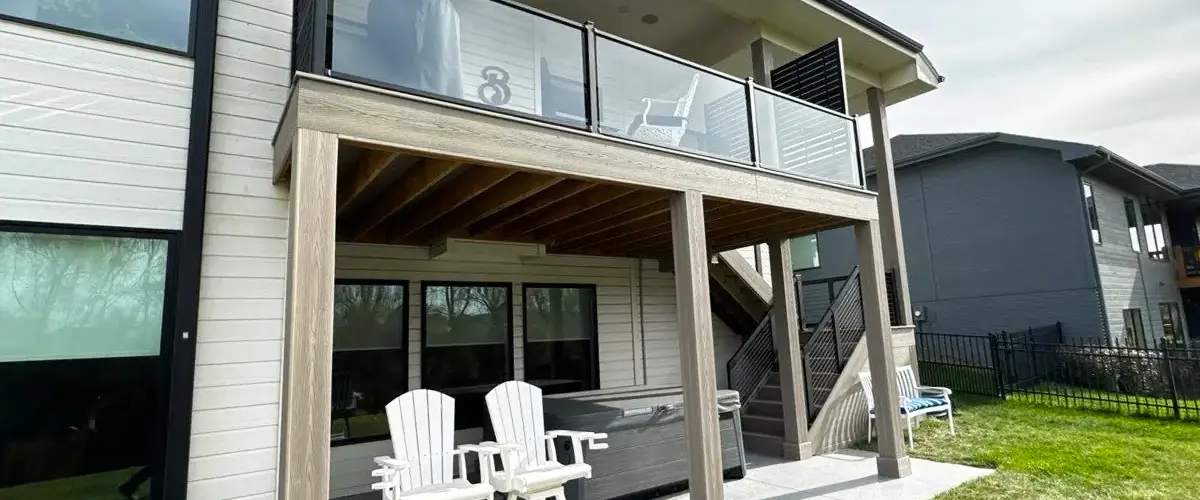 Modern two-story home with elevated covered deck, glass railings, and white bar stools on a finished concrete patio.