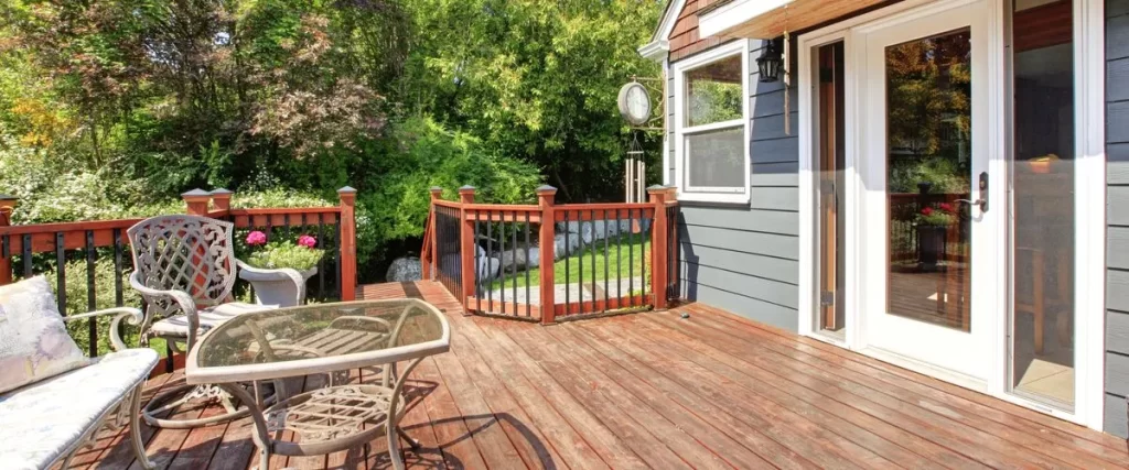 Cozy wooden deck featuring dark wrought iron patio chairs and a glass-top table, enclosed by a short wooden railing, next to a home with gray siding and white sliding glass doors. Lush green foliage and trees are visible in the background.