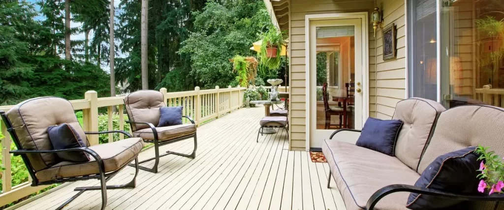 Large outdoor deck painted in a light beige/cream color, furnished with matching tan patio chairs and a loveseat, overlooking a lush green forest backdrop.