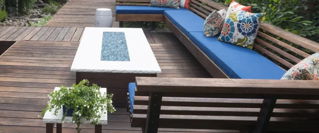 Dark brown wooden deck with long built-in benches featuring vibrant blue cushions, a white coffee table with a blue glass fire feature, and a small potted plant.