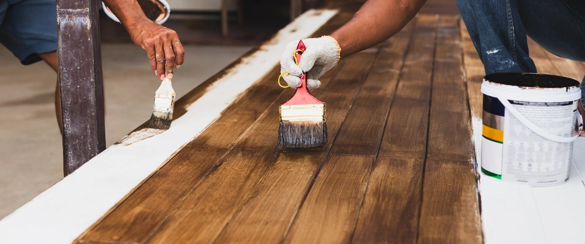Two professional workers applying dark brown wood stain to deck boards with brushes next to a white painted border.