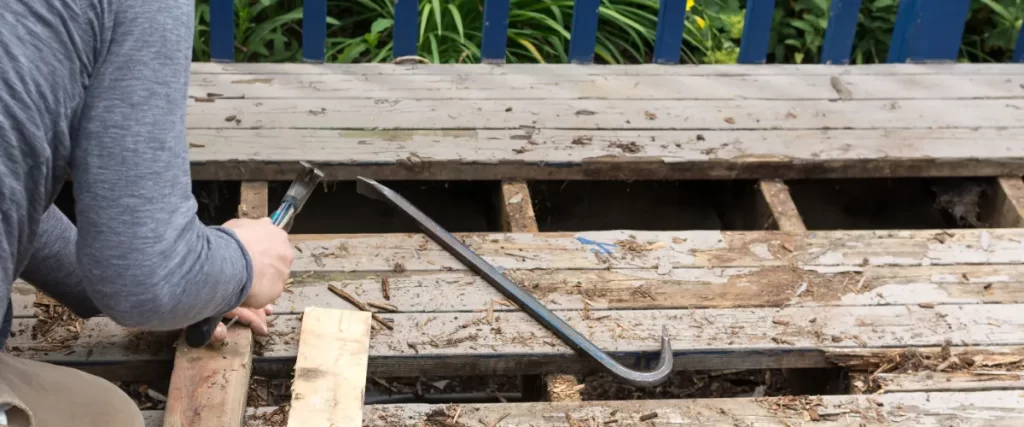A person is using a hammer and a pry bar to remove old, weathered wooden deck boards, exposing the underlying joists