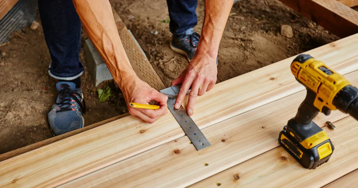 a person using a speed square and pencil to mark a wooden deck board during construction
