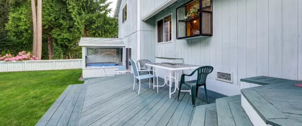 a wooden deck attached to the back of a light blue house, featuring outdoor furniture and a small covered area with a hot tub
