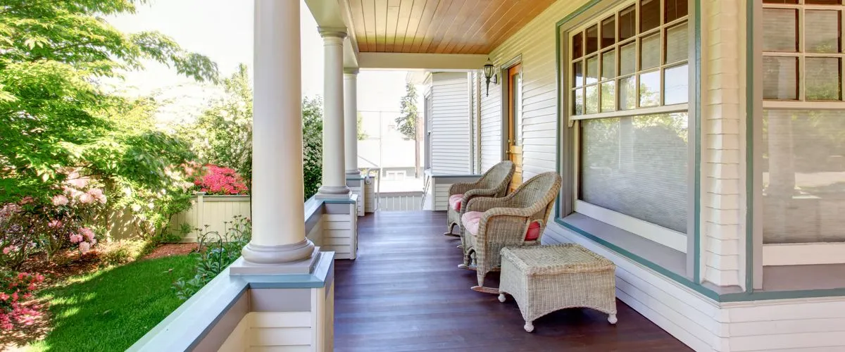 Deck Replacement A covered craftsman style front porch featuring dark stained wood floorboards, white architectural columns, and a pair of wicker rocking chairs with pink cushions overlooking a lush green garden with blooming pink rhododendrons.