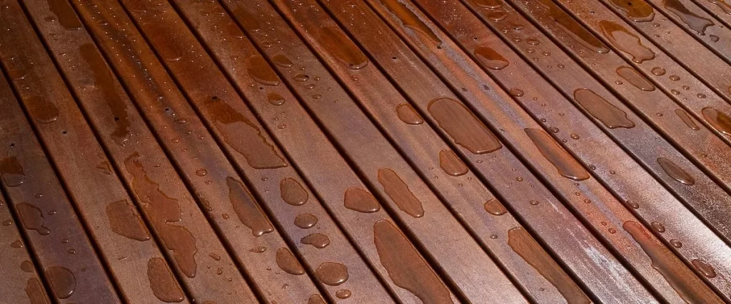 A detailed close-up view of a waterproof deck showing rainwater beading up on the surface of rich, reddish-brown wooden planks, demonstrating the effectiveness of water-repellent wood sealant and stain.