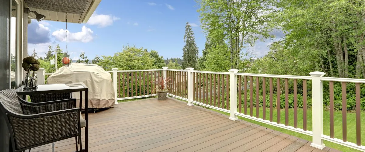 A beautifully remodeled outdoor deck featuring wide, brown composite decking and a white vinyl railing with contrasting brown balusters, overlooking a lush green yard and tall trees.