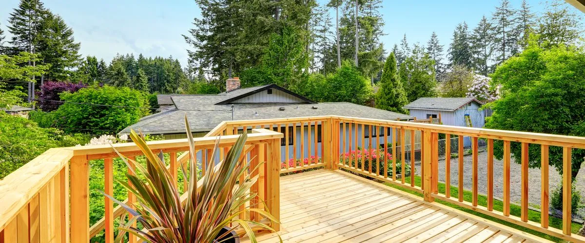 A wide-angle view of a professionally constructed elevated wooden deck made of light-colored cedar, featuring a sturdy vertical slat railing system overlooking a lush green backyard with tall evergreen trees and a neighboring house.