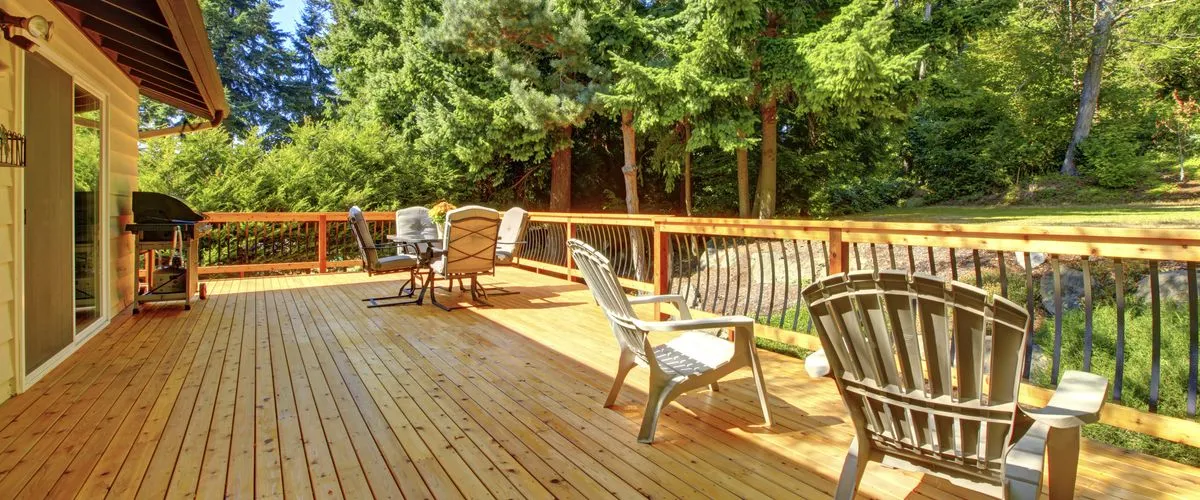 A spacious light brown cedar wood deck featuring a dining set, white Adirondack chairs, a barbecue grill, and modern black curved metal railings with a scenic view of a lush evergreen forest.