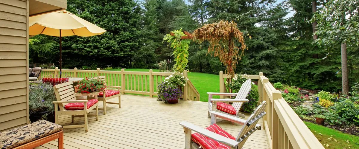 A spacious, light-colored wooden backyard deck featuring a matching railing, comfortable wooden lounge chairs with red cushions, a patio umbrella, and a scenic view of a lush green lawn and evergreen forest.