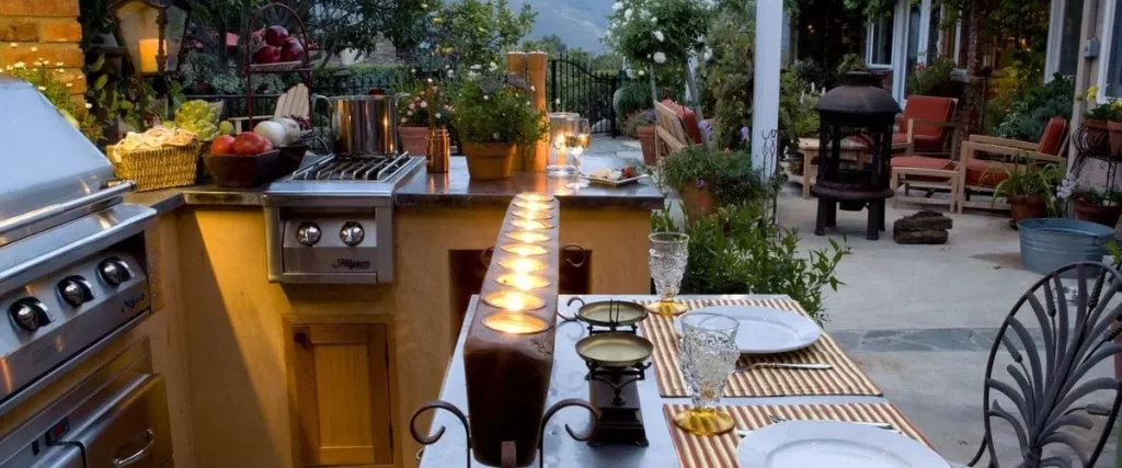 Outdoor Kitchens On Decks In Omaha, NE Luxury outdoor kitchen and dining patio at night, featuring a stainless steel grill, built-in gas range, counter with fresh produce, and a set table with wine glasses and a large candle centerpiece.