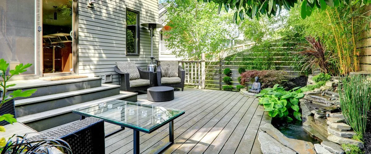 A modern gray multi-level composite deck featuring a built-in stone waterfall pond, black wicker patio furniture with plush cushions, a glass-top coffee table, and lush green landscaping with bamboo privacy screening.