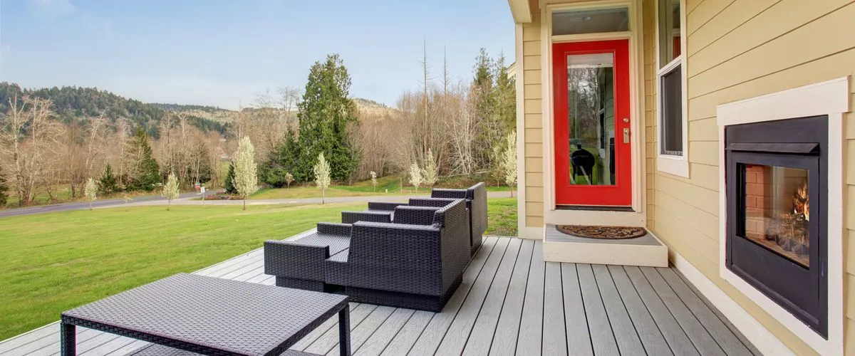 A modern gray composite wood deck featuring black wicker lounge chairs and a matching coffee table, situated next to a tan house with a vibrant red door and an outdoor built-in fireplace overlooking a green valley.