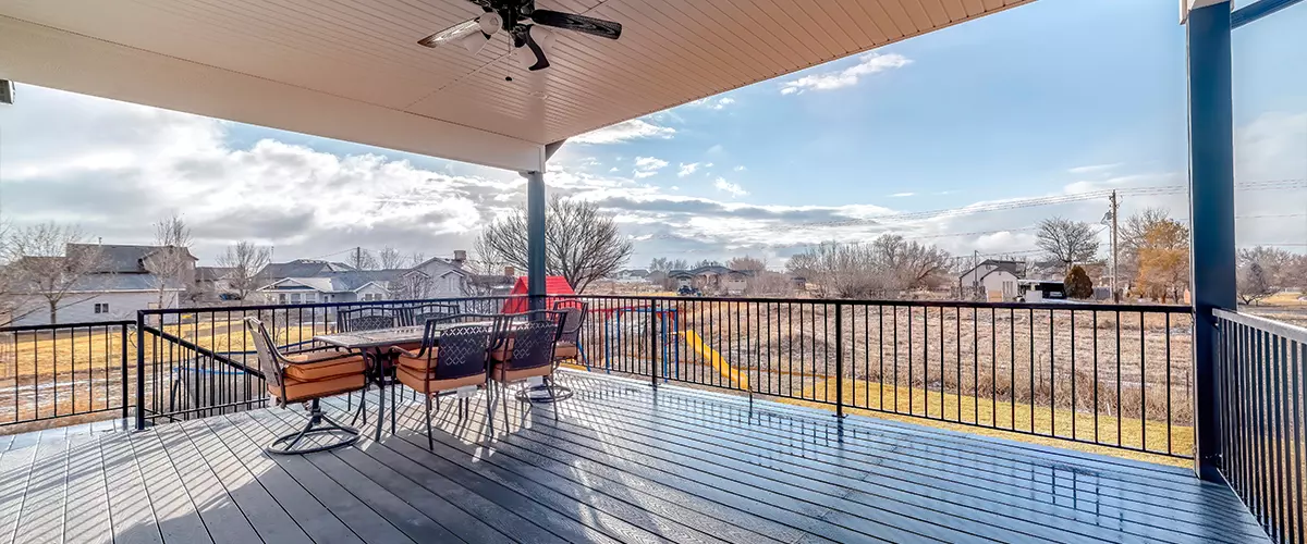 A spacious second-story covered deck featuring dark grey composite flooring, a black metal safety railing, and a patio dining table with orange cushioned chairs overlooking a residential neighborhood.