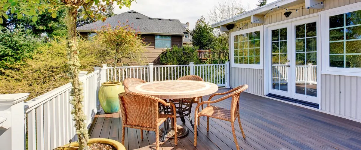 A spacious dark wood outdoor deck featuring a round tile-top table surrounded by four brown wicker chairs, enclosed by white classic railings with large potted plants and a view of lush green trees and neighboring houses.