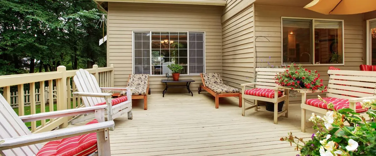 Spacious beige wooden outdoor deck featuring white Adirondack chairs with red striped cushions, wooden lounge chairs, and potted flowers against tan house siding and a lush green forest background.
