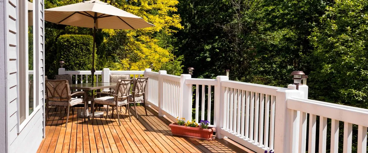 Deck Building In Louisville, NE A sunlit outdoor wooden deck made of natural cedar planks featuring a white vinyl safety railing, a patio dining table with four cushioned chairs, and a large tan umbrella surrounded by lush green trees.