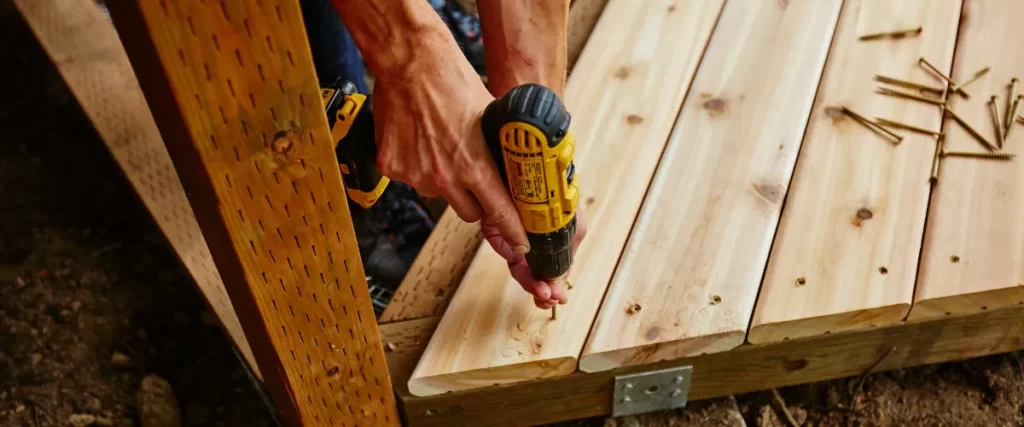 a close-up of a person using a power drill to fasten a wooden deck board with a screw
