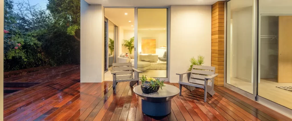 Smart Tech Upgrades for Modern Decks in 2026 (Lighting, Outlets, Sound) a modern patio area with a wooden deck and large sliding glass doors leading into an illuminated interior living space