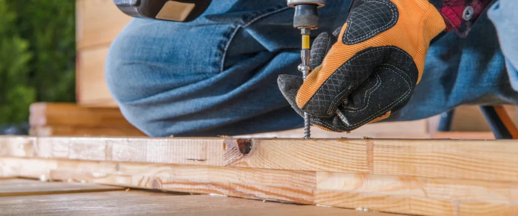 Wind-Resistant Deck Design for the Omaha–Lincoln Corridor A worker wearing protective gloves using a power drill to drive a stainless steel deck screw into a pressure-treated wood board.
